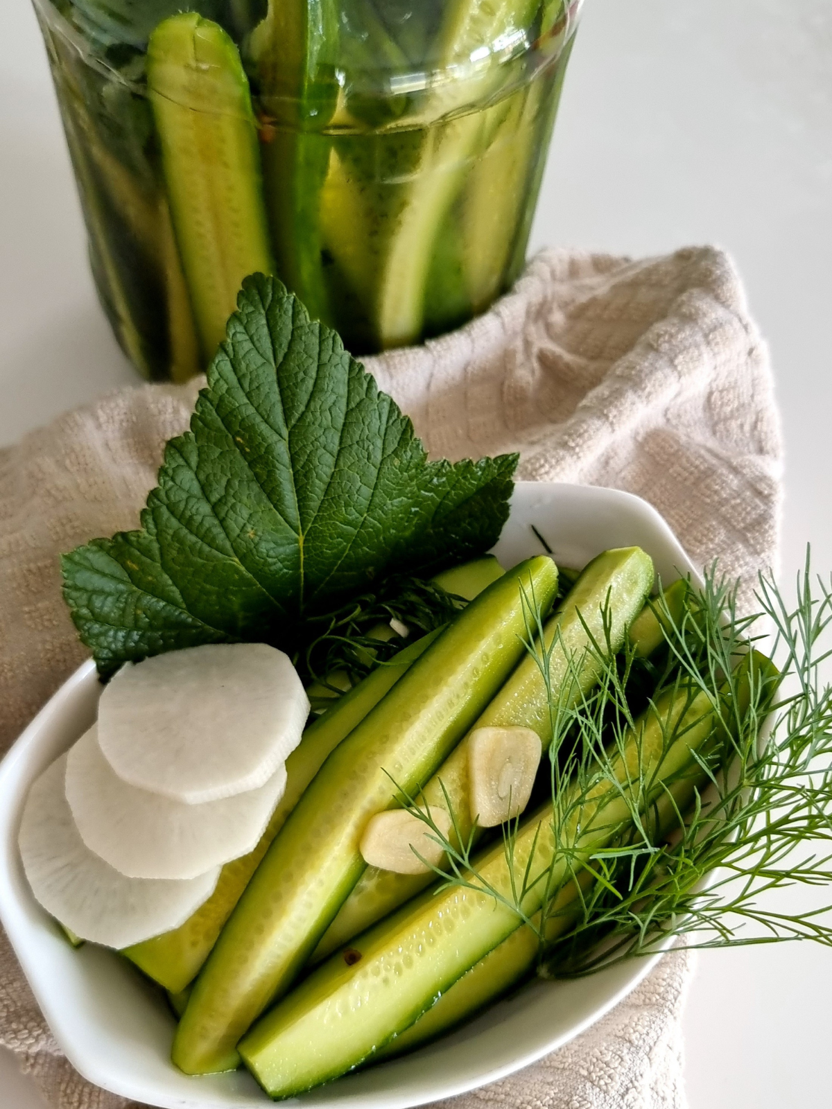 Frische Malosolnye Gurken mit Dill, Knoblauch und geschnittenen Gurkensticks in einer weißen Schale auf hellem Leinentuch. Im Hintergrund steht ein Glas mit eingelegten Gurken in natürlicher Küchenatmosphäre. Das Bild zeigt eine traditionelle, leicht gesalzene Gurkenzubereitung mit frischen Kräutern und sommerlichem Charakter.