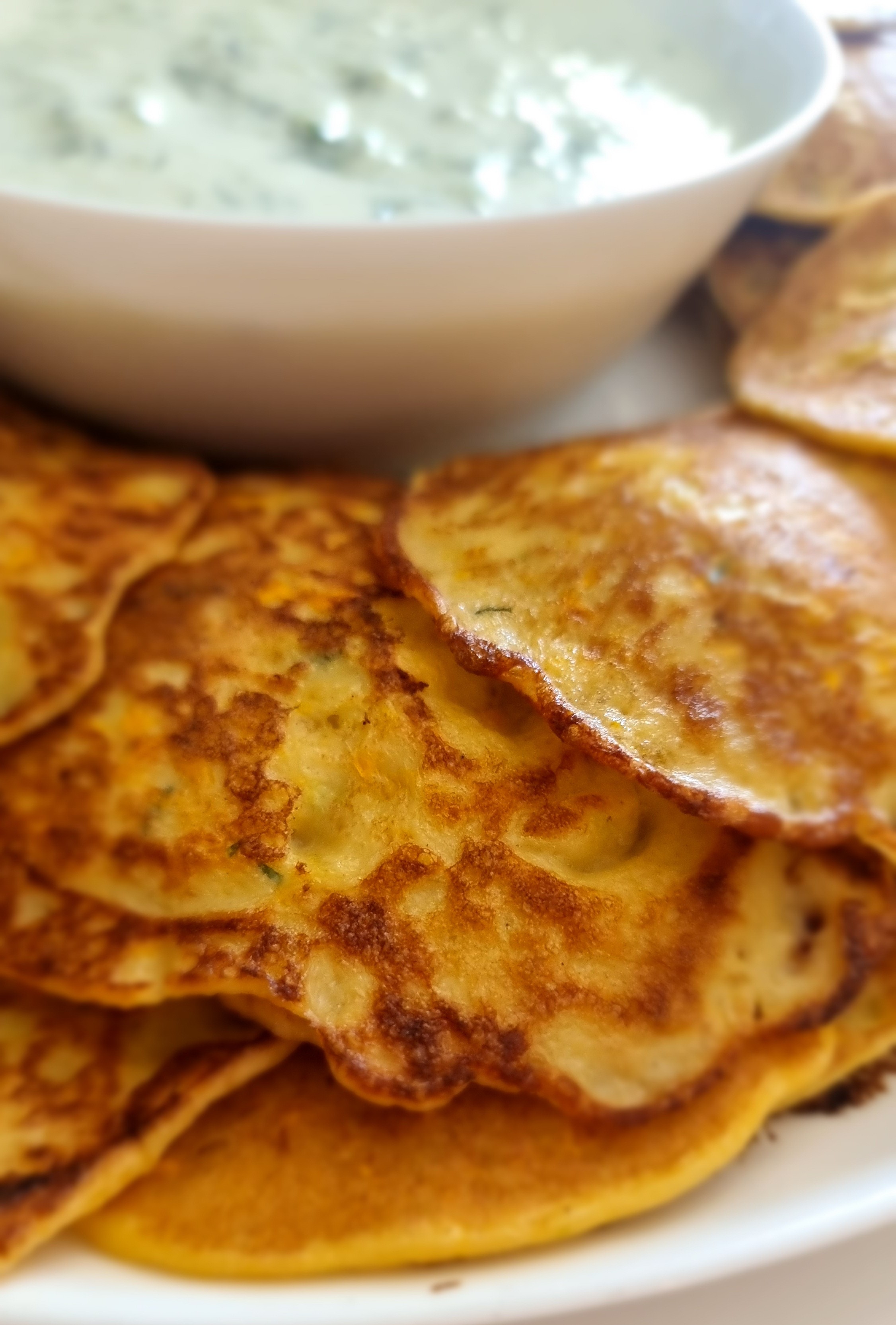 Schmackhafte gebratene Zucchinipuffer auf einem Teller mit Schafskäse-Dip im Hintergrund.