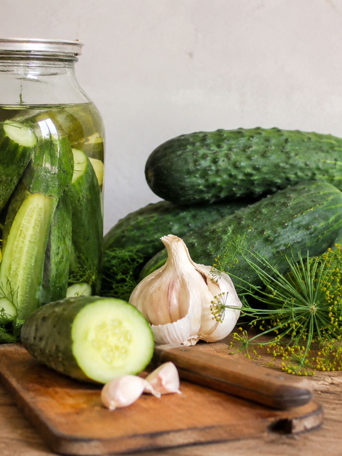 Selbstgemachten Essiggurken im Glas auf einem Holztisch. Frische Gurken, Dillblüten und Knoblauch liegen daneben in natürlichem Licht. Das Bild zeigt ein einfaches Rezept mit Apfelessig und sommerlichen Gartenkräutern in gemütlicher Küchenatmosphäre.